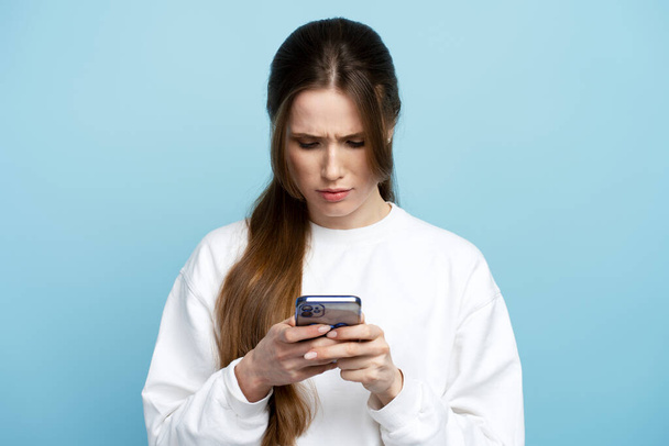 Poważna, zamyślona kobieta w białej koszuli trzymająca smartfona w rękach, rozmawiająca z przyjaciółmi lub ustalająca porządek, mająca skoncentrowany wyraz twarzy. Kryty studio strzał izolowany na niebieskim tle  - Zdjęcie, obraz