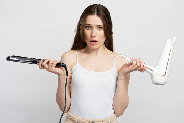 Beautiful surprised woman holding hair straightener and hair dryer isolated on white background. Beauty, morning routine, hair care concept  - Valokuva, kuva