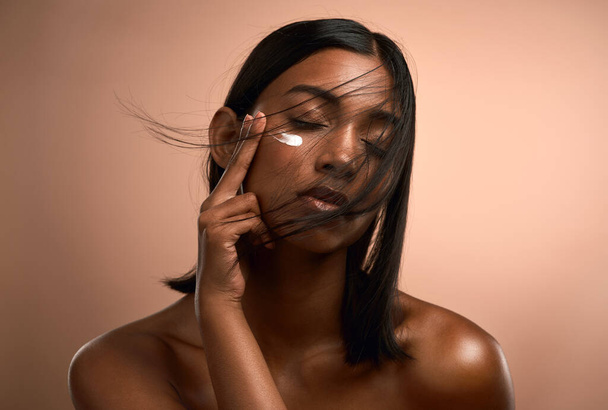 Beauty need only be a whisper. an attractive young woman applying moisturiser against a brown background - Foto, afbeelding
