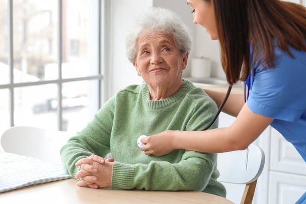 Junge Pflegerin hört Seniorin mit Stethoskop in Küche - Foto, Bild