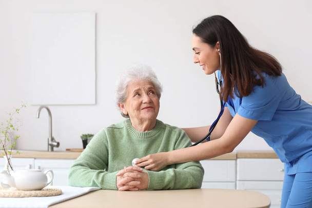 Junge Pflegerin hört Seniorin mit Stethoskop in Küche - Foto, Bild