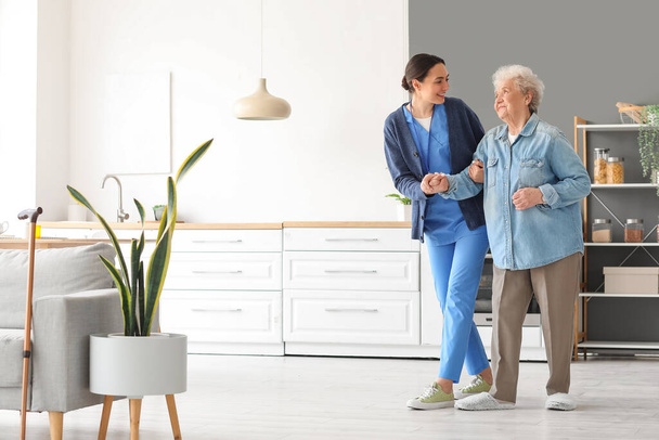 Young caregiver helping senior woman to walk in kitchen - Foto, afbeelding