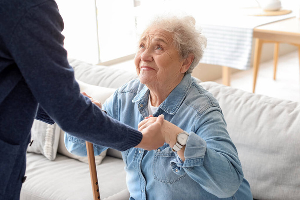 Νεαρός φροντιστής βοηθά ηλικιωμένη γυναίκα με ραβδί για να σταθεί στο σπίτι - Φωτογραφία, εικόνα