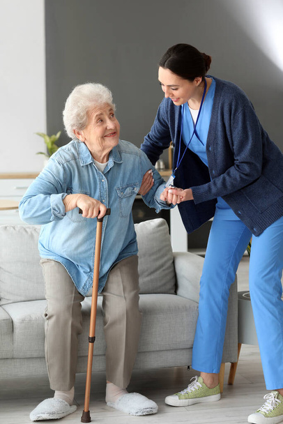 Young caregiver helping senior woman with stick to stand up at home - Фото, изображение