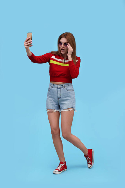 Beautiful woman in sunglasses taking selfie on light blue background - Фото, зображення