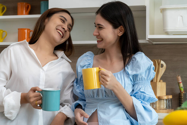 Happy Asian young pretty woman and her girlfriend holding coffee mugs and chatting together in kitchen, The moment romantic time of female lover, LGBTQ freedom of love concept, copy space - Foto, Imagen