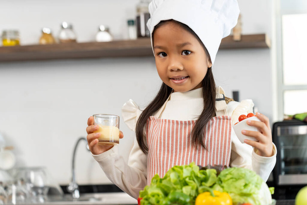 Portrait lovely girl holding glass of condiment and red tomato in white bowl in hand, smile and looking at camera, Vegetables, bell peppers, eggs and cooking utensils on counter in kitchen, copy space - Photo, image