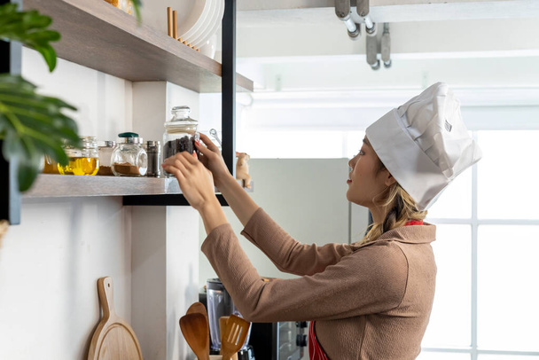 Beautiful woman in chef's hat and apron cleaning and organizing utensils on kitchen shelf at home, copy space - Foto, Bild