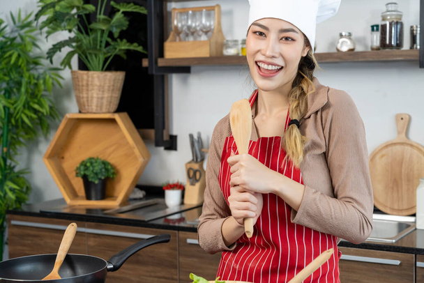 Portrait Asian pretty woman in chef hat and apron hold a wooden spade of frying pan in kitchen, smile and looking at camera, copy space - Foto, Bild