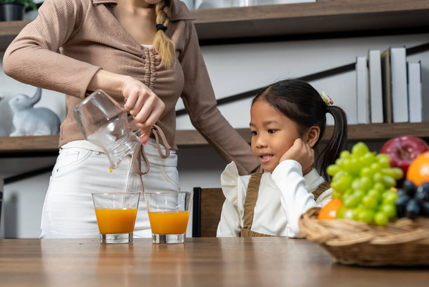 Hermosa madre vertiendo jugo de naranja fresca en el vaso para su encantadora hija en la mesa en la cocina de la casa, que hablan y sonríen con feliz juntos, espacio de copia - Foto, Imagen