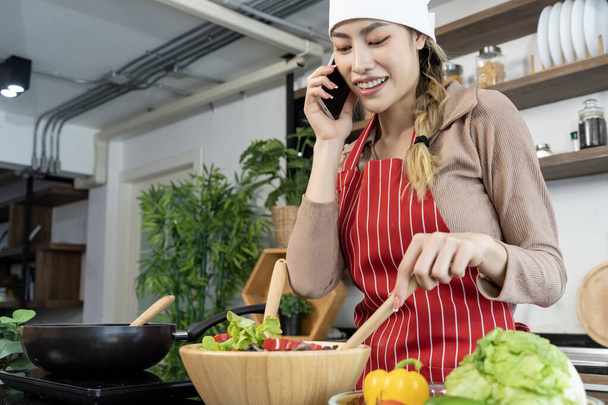 Portrait Asian woman in chef hat and apron talking with smartphone while cooking salad vegetables in wooden bowl, pan with electric stove eggs place on counter in kitchen, copy space - Φωτογραφία, εικόνα