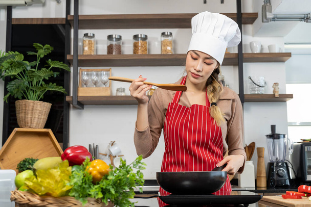 Portrait Asian woman in chef hat and apron cooking in pan with electric stove in kitchen, , eggs, vegetable and kitchen equipment place on counter, copy space - Foto, Imagem