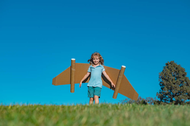 Child boy plays in an astronaut dreams of space. Happy child play with toy plane cardboard wings against blue sky. Kid having fun in summer field outdoor. Portrait of boy with paper wings - Photo, Image