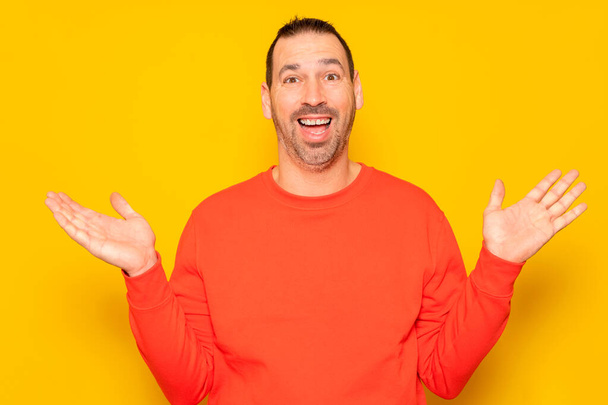 Hombre sonriente divertido en suéter rojo mostrando las palmas hacia arriba, gesto sorpresa inesperada aislado sobre fondo amarillo. Atractivo hombre feliz escuchando buenas noticias, la captura de algo - Foto, imagen