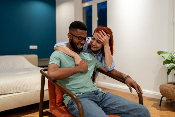 Überglückliche Paare unterhalten sich entspannt am Familienabend im neuen Mietwohnsitz. Lächelnder Afroamerikaner genießt auf Sessel Kommunikation mit umarmenden europäischen lachenden Frau Freundin - Foto, Bild