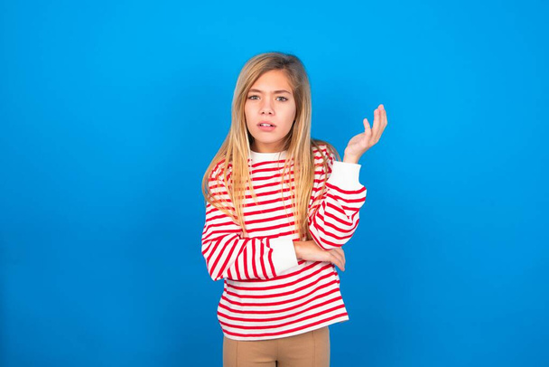 Studio shot of frustrated teen girl wearing striped shirt over blue background gesturing with raised palm, frowning, being displeased and confused with dumb question. - Foto, immagini