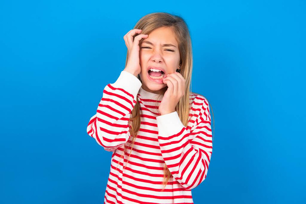 Young gloomy beautiful caucasian teen girl wearing striped shirt over blue background, hiding face with hands pouting and crying, standing upset and depressed complaining about job problem. - Photo, Image