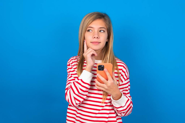 Thinking dreaming teen girl wearing striped shirt over blue background using mobile phone and holding hand on face. Taking decisions and social media concept. - Photo, Image