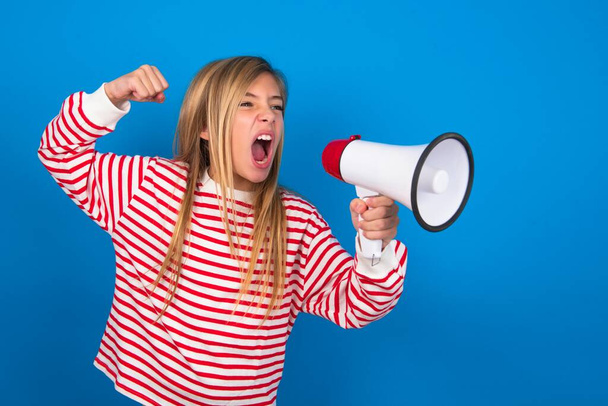 dospívající dívka v pruhované košili přes modré pozadí komunikuje křičí hlasitě drží megafon, vyjadřuje úspěch a pozitivní koncept, nápad pro marketing nebo prodej. - Fotografie, Obrázek