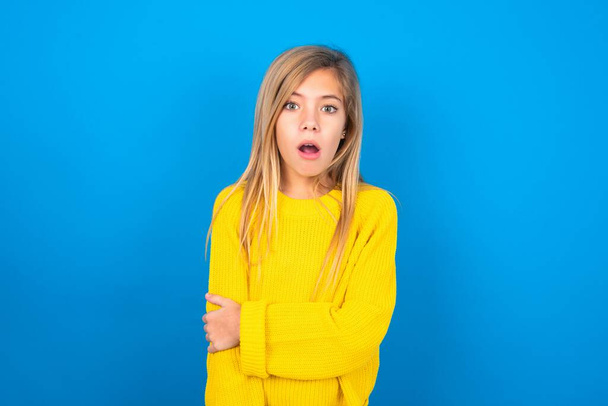 Shocked embarrassed caucasian teen girl wearing yellow sweater over blue studio background keeps mouth widely opened. Hears unbelievable novelty stares in stupor - Valokuva, kuva
