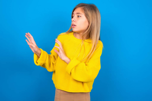 Dispiaciuto caucasico teen girl indossa maglione giallo su sfondo blu studio mantiene le mani verso lo spazio vuoto e chiede di non avvicinarsi vede qualcosa di spiacevole - Foto, immagini