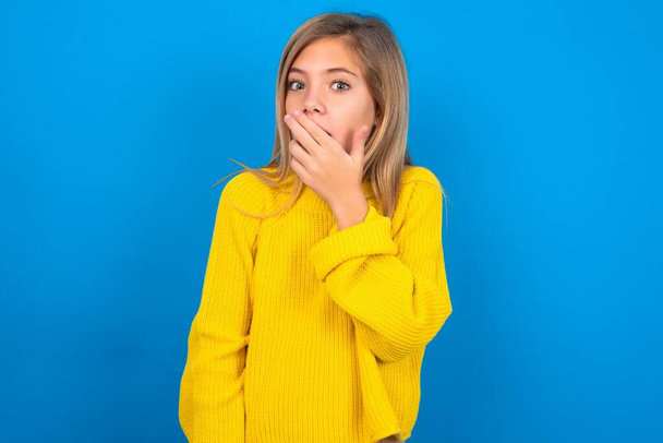 Blondýny kavkazský dospívající dívka na sobě žlutý svetr přes modrý studio pozadí zakrývá ústa a dívá se s úžasem na kameru, nemůže uvěřit neočekávané fámy.  - Fotografie, Obrázek