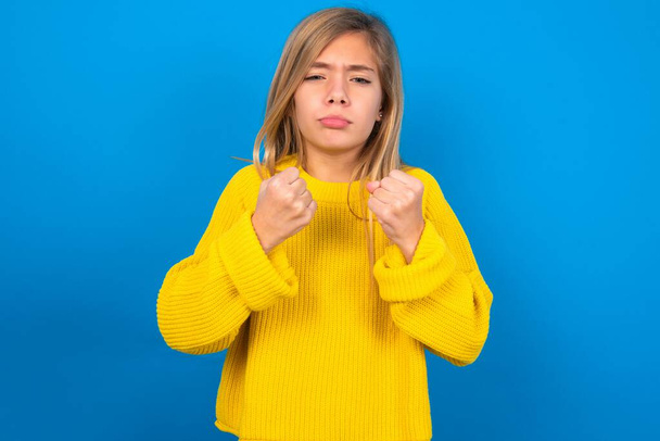 Displeased annoyed caucasian teen girl wearing yellow sweater over blue studio background clenches fists, gestures pissed, ready to revenge, looks with aggression at camera stands full of hate, being pressured  - Фото, зображення