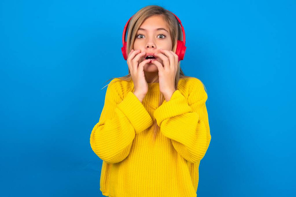 Šokovaný kavkazský dospívající dívka na sobě žlutý svetr přes modré studio pozadí zírá ustrašený na kameru udržuje ústa široce otevřené nosí bezdrátové stereo sluchátka na uších - Fotografie, Obrázek