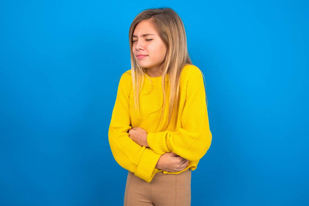 Caucasian teen girl wearing yellow sweater over blue studio background has stomachache  - Photo, Image