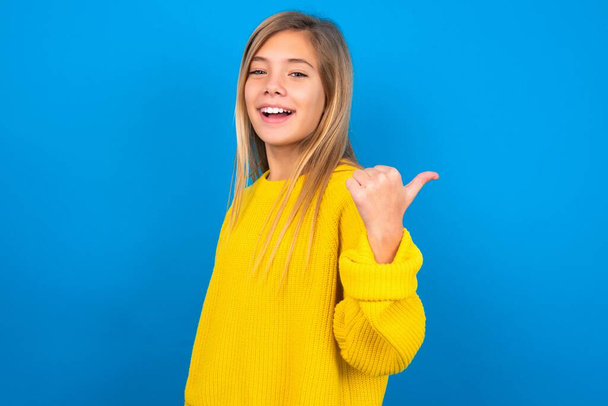 Impressed caucasian teen girl wearing yellow sweater over blue studio background points back empty space - Zdjęcie, obraz