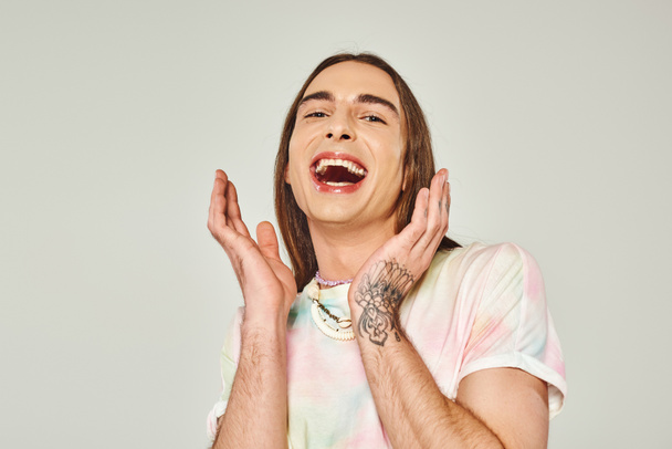 tattooed and excited young gay man with long hair and opened mouth looking at camera and gesturing with hands on grey background, pride month concept - Photo, Image