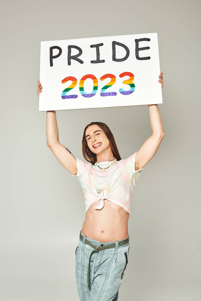 cheerful young gay man with tattoo and long hair standing in denim jeans and tied knot on t-shirt showing his belly and holding pride 2023 placard on grey background  - Foto, imagen