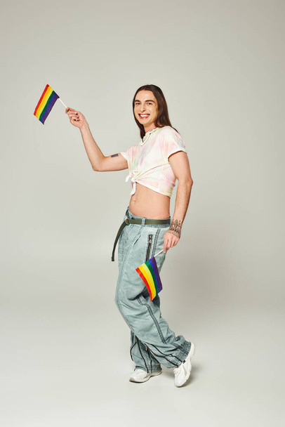 cheerful and tattooed gay man with long hair and bare belly standing in denim jeans while holding rainbow flags for pride month on grey background  - Photo, Image