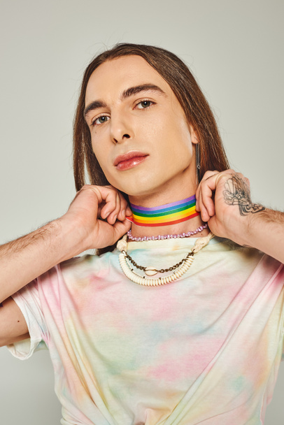 tattooed gay man with long hair and tie dye t-shirt holding rainbow lgbt flag near neck during pride month and looking at camera on grey background  - Foto, Bild