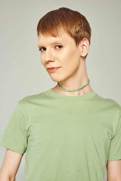 retrato de una joven persona no binaria con brillo labial brillante de pie en camiseta verde y mirando a la cámara durante el mes de orgullo sobre fondo gris - Foto, Imagen