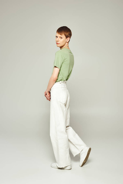 full length of young queer person with lip gloss standing in green t-shirt and white denim jeans while looking at camera on pride month on grey background - 写真・画像