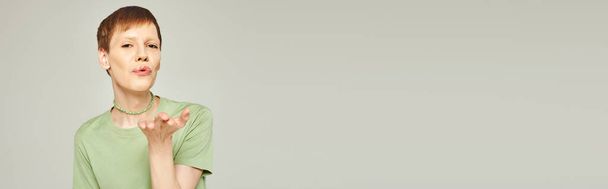 joven queer persona con brillo de labios de pie en verde camiseta y soplado aire beso mientras mira a la cámara durante el mes de orgullo sobre fondo gris, pancarta  - Foto, Imagen