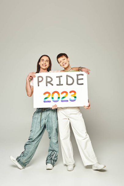 happy lgbt friends holding pride 2023 placard and looking at camera while celebrating lgbtq community holiday in June on grey background in studio  - Foto, Bild