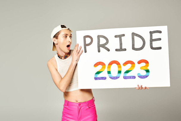 tetovaný a šokovaný gayové v baseballové čepici, tričku a růžové kalhoty drží hrdost 2023 plakát, zatímco stojí s otevřenými ústy na šedém pozadí  - Fotografie, Obrázek