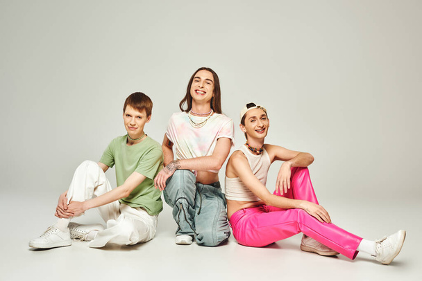 Diverso grupo de personas de la comunidad lgbtq felices y jóvenes en ropa colorida sonriendo mientras se sientan juntos y celebran el mes del orgullo sobre fondo gris en el estudio  - Foto, Imagen