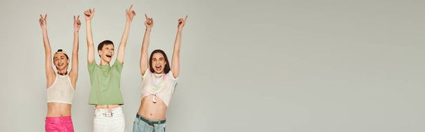 happy and young lgbt community friends in colorful clothes smiling while looking at camera, raising hands and celebrating pride month together on grey background in studio, banner  - Photo, Image