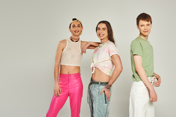 diverse and young lgbtq community friends with tattoos standing in colorful clothes smiling while looking at camera in studio on pride month, grey background  - Photo, Image