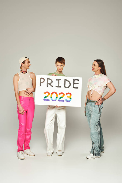 happy gay men in colorful clothes standing and looking at queer friend holding pride 2023 placard while celebrating lgbt community holiday in June, grey background, studio  - Foto, afbeelding