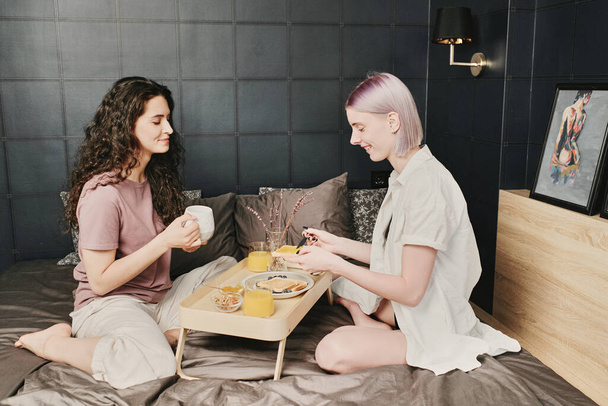 sourire caucasien gay couple assis sur lit et boire café tout en mangeant pain grillé pour petit déjeuner - Photo, image