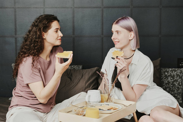 Positive young Caucasian lesbian couple sitting on bed with tray and eating toasts in morning - Фото, зображення