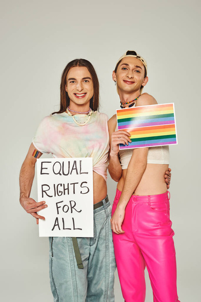 szczęśliwi i wytatuowani przyjaciele lgbt trzymający tęczową flagę na zdjęciu i tabliczce z równymi prawami dla wszystkich liter stojąc razem w dzień dumy, szare tło  - Zdjęcie, obraz