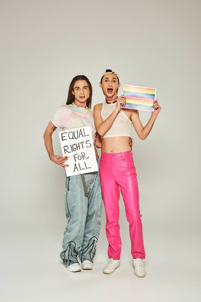 emotional and tattooed lgbt people holding rainbow flag picture and placard with equal rights for all lettering while standing together on pride day, grey background  - Foto, imagen