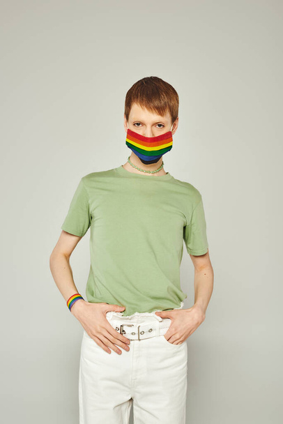 portrait de jeune queer debout en t-shirt vert et masque de drapeau lgbt tout en regardant la caméra et posant pendant le mois de la fierté sur fond gris - Photo, image