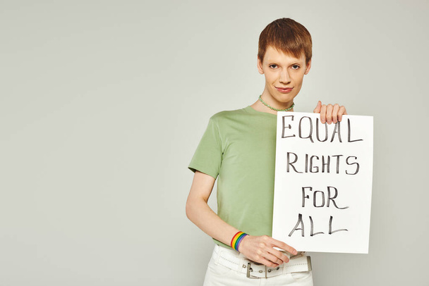Porträt einer jungen queeren Aktivistin mit lgbt-Fahnenarmband und gleichberechtigtem Plakat für alle Schriftzüge, die während eines stolzen Monats auf grauem Hintergrund in die Kamera blickt - Foto, Bild