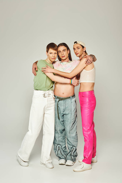 täyspitkä kolme lgbtq ihmistä värikkäissä vaatteissa katsoen kameraa ja halaten toisiaan harmaalla taustalla studiossa, ylpeyden kuukauden konseptin juhliminen   - Valokuva, kuva